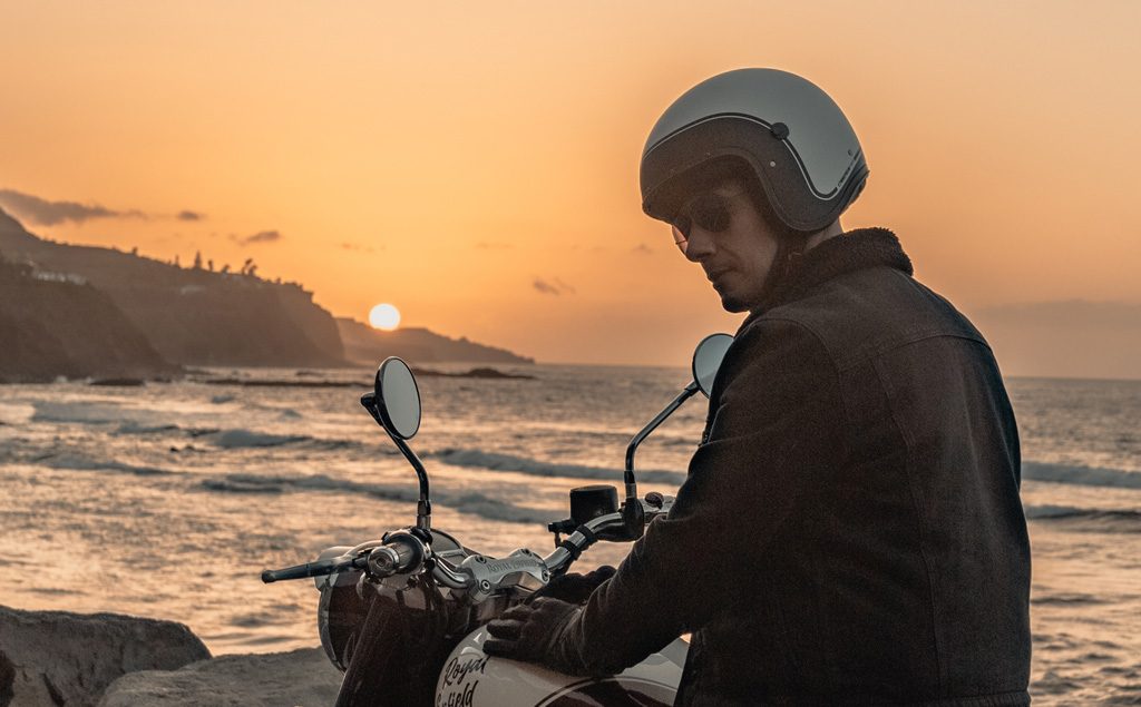 uomo in sella alla sua moto in riva al mare al tramonto, indossando un casco in stile vintage di Caberg